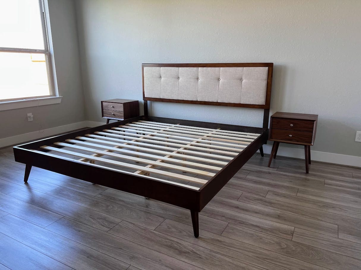 Wooden bed frame with tufted headboard in a room with wooden flooring and a window.
