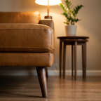A close-up of a tan leather armchair in a warm, modern living room setting with a lit floor lamp and a small side table holding a potted plant.
