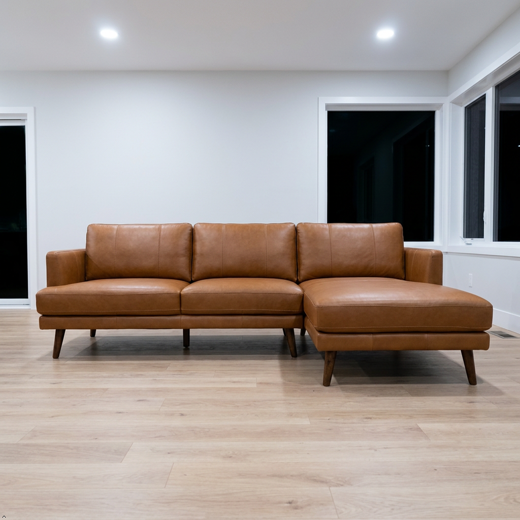 A modern L-shaped sectional sofa in tan leather with dark wooden legs, positioned on a light wood floor in a minimalist room with white walls and windows.