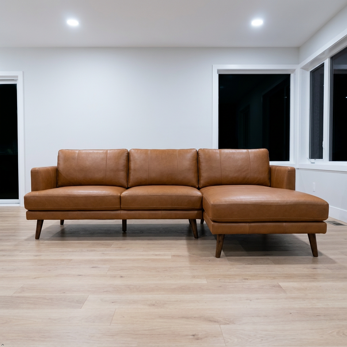 A modern L-shaped sectional sofa in tan leather with dark wooden legs, positioned on a light wood floor in a minimalist room with white walls and windows.