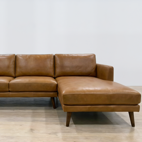 A mid-century modern three-seat leather sofa with a right-facing chaise lounge, upholstered in warm cognac brown leather with dark tapered wooden legs, set against a minimalist white wall and light wood floor.