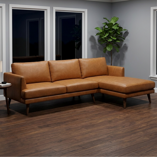 A modern tan leather sectional sofa with a right-facing chaise lounge, positioned in a contemporary living room with gray walls, dark wood floors, a side table with a lamp, and a potted plant.