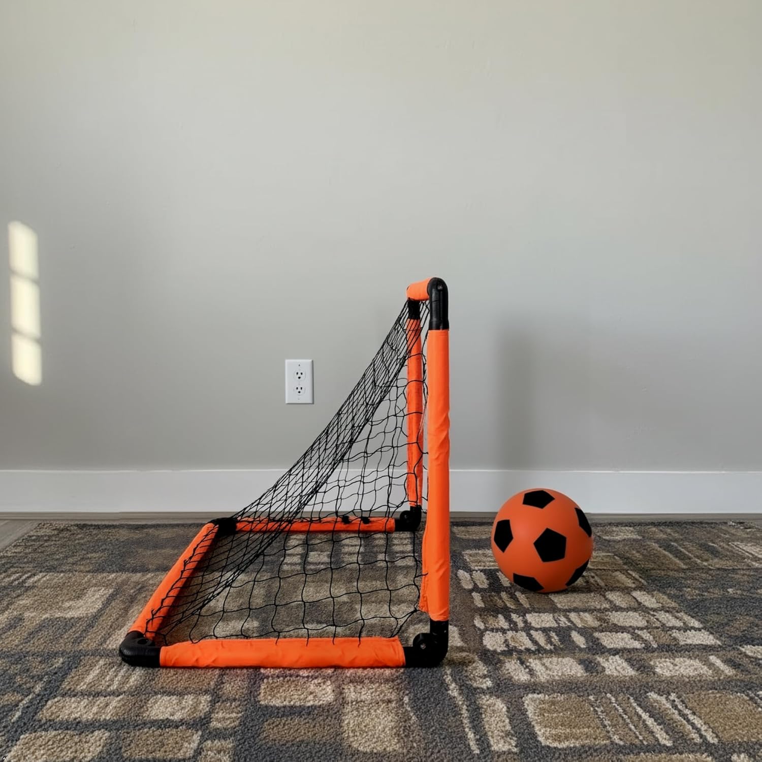 Mini soccer goal with orange frame and black net on a carpeted floor against a gray wall.