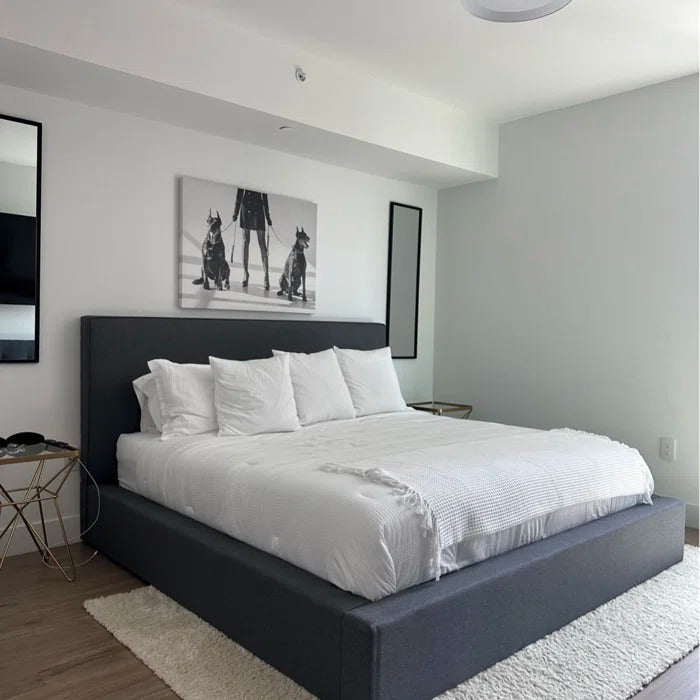 A modern platform bed with a dark gray upholstered frame and white bedding, styled in a minimalist bedroom setting with a black and white dog art print on the wall.