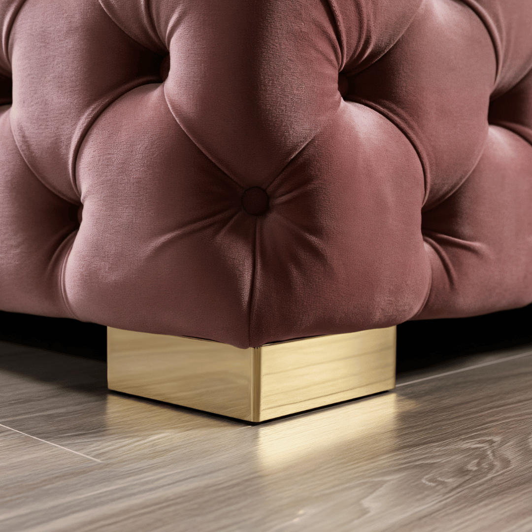 Close-up of a pink tufted ottoman with a gold base on a wooden floor.