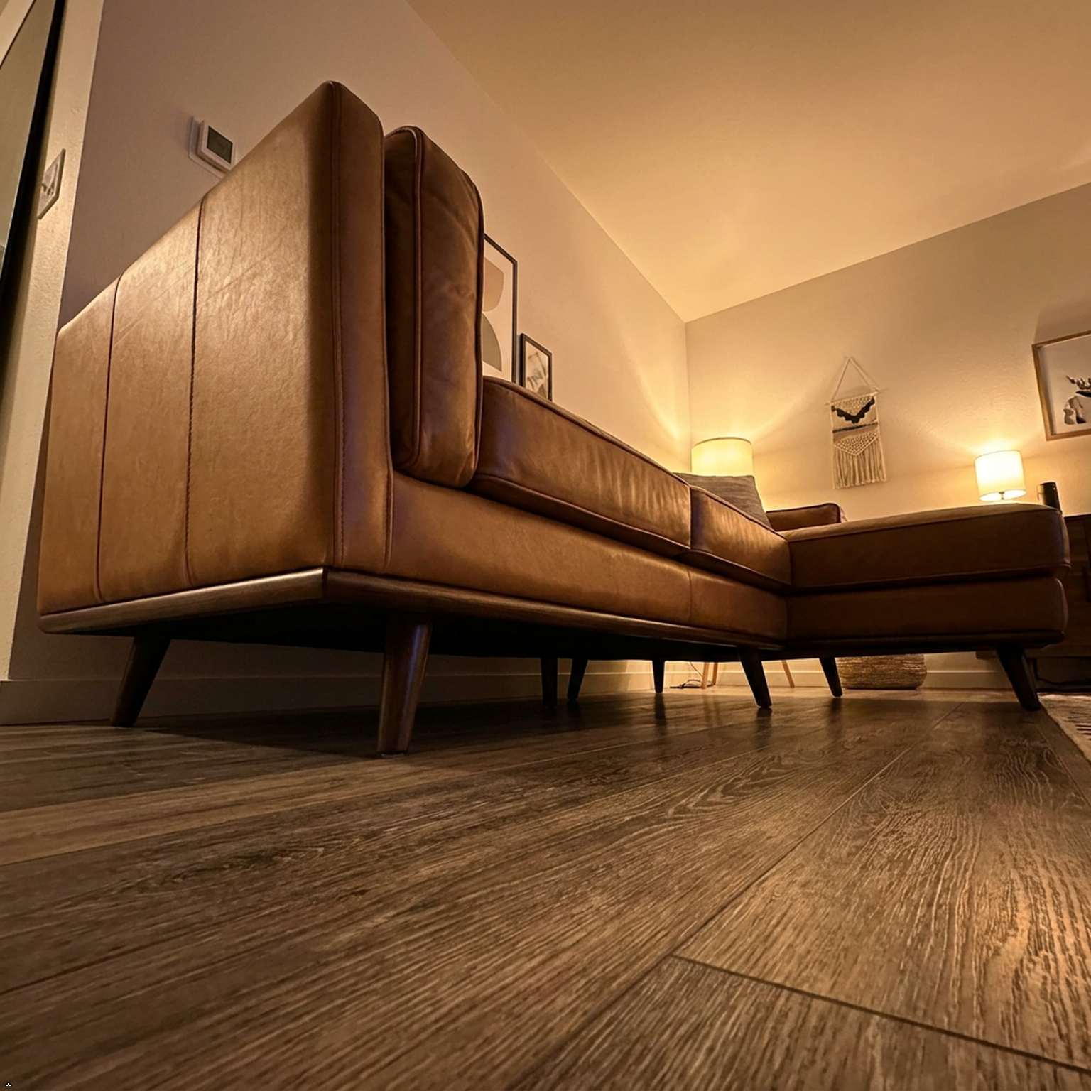 A low-angle shot of a modern brown leather sectional sofa with a chaise lounge, set in a cozy living room with wood floors and warm ambient lighting.
