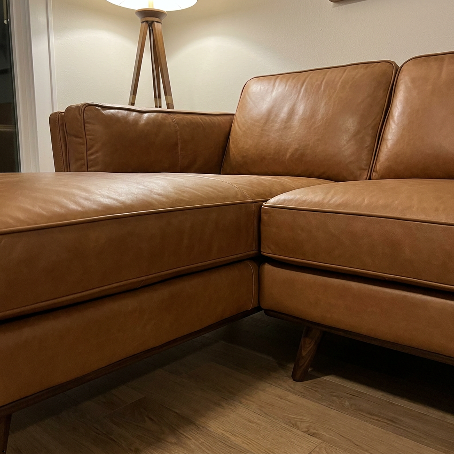 A close-up of a modern sectional sofa corner upholstered in warm tan leather, showing the seat cushion, back cushions, and dark wood legs on a wood floor.