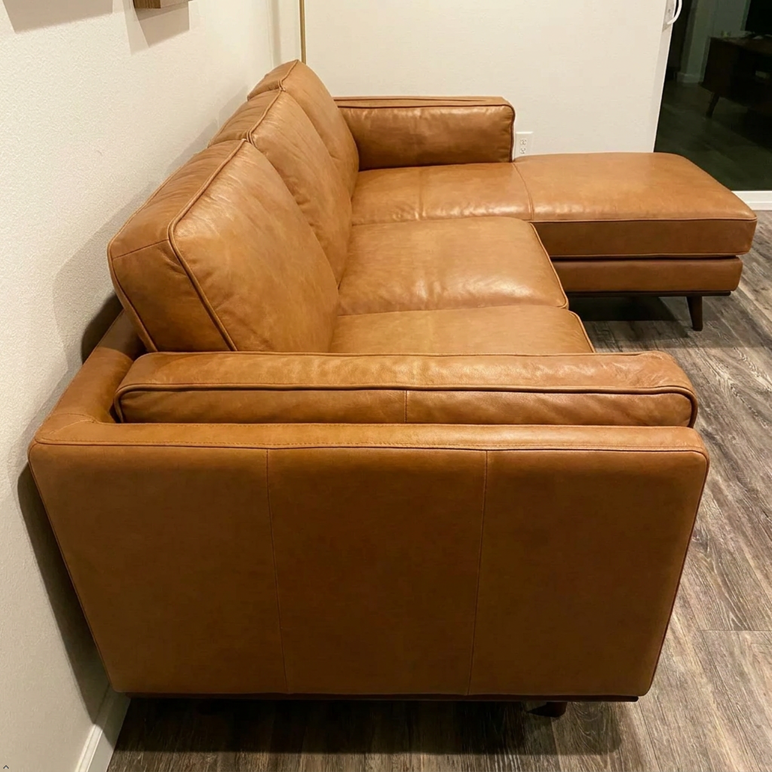 A tan leather sectional sofa with a right-facing chaise lounge, photographed in a home setting on a gray wood-look floor against a white wall.