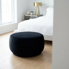 Black ottoman in a bedroom setting with a bed and lamp in the background.