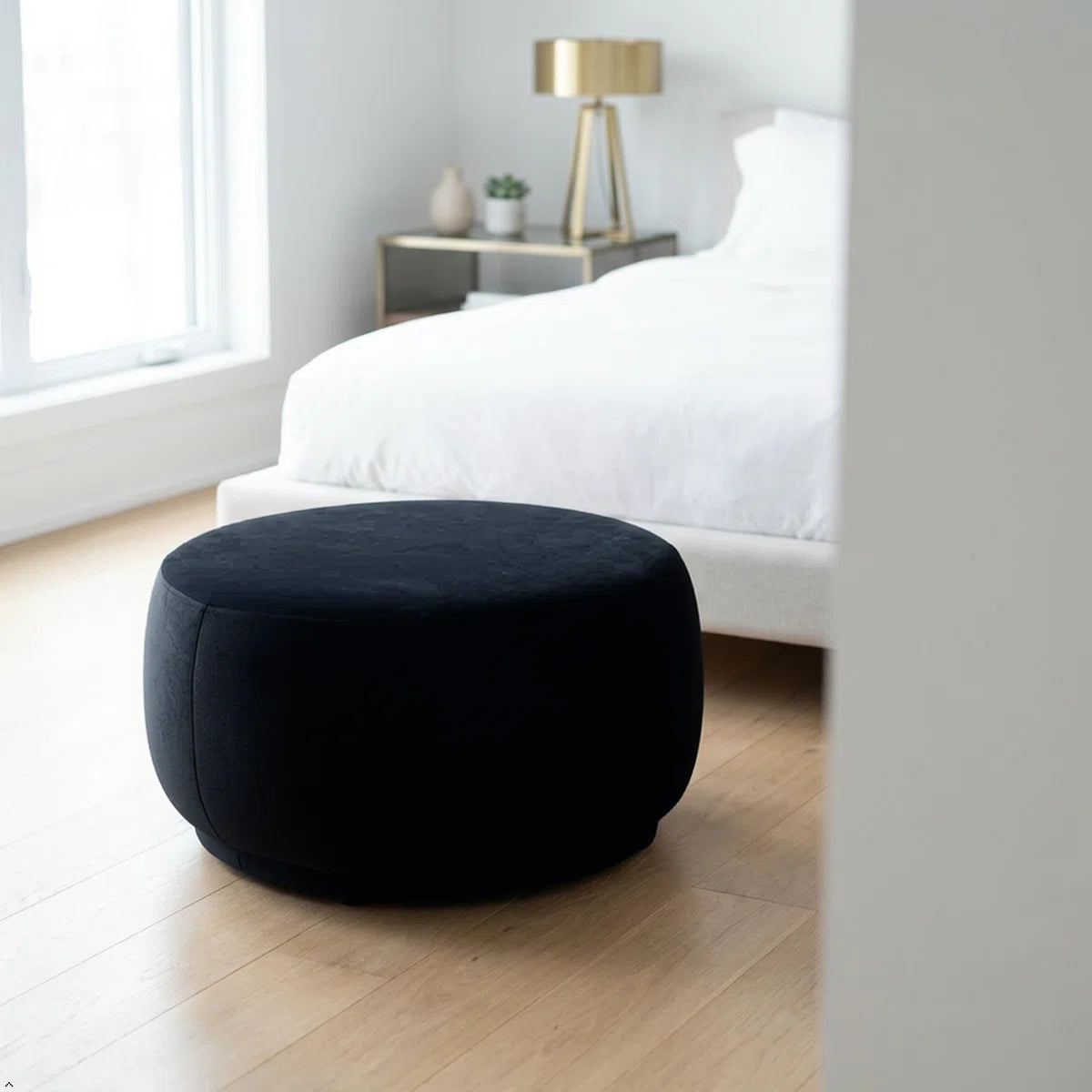 Black ottoman in a bedroom setting with a bed and lamp in the background.