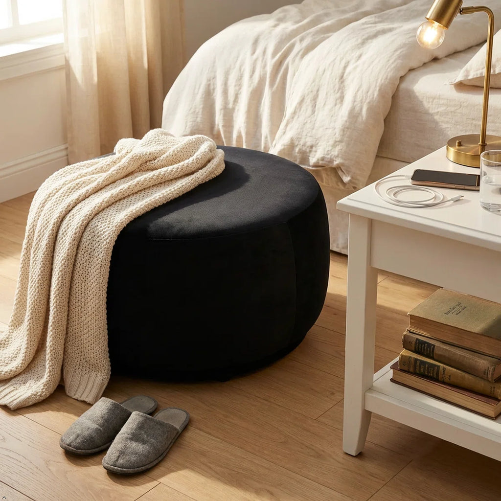 Black ottoman with a blanket, gray slippers, and a nightstand with books in a bedroom.