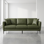 An olive green leather three-seater sofa with two matching throw pillows, set in a minimalist room with a concrete floor and white walls.