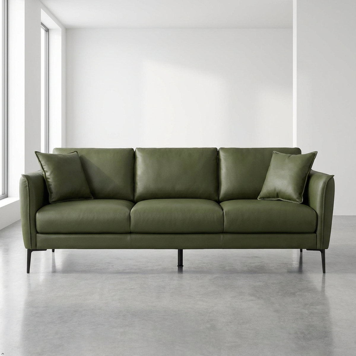 An olive green leather three-seater sofa with two matching throw pillows, set in a minimalist room with a concrete floor and white walls.