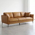 A mid-century modern three-seater sofa upholstered in warm tan leather, shown in a minimalist studio setting with a concrete floor and white walls.