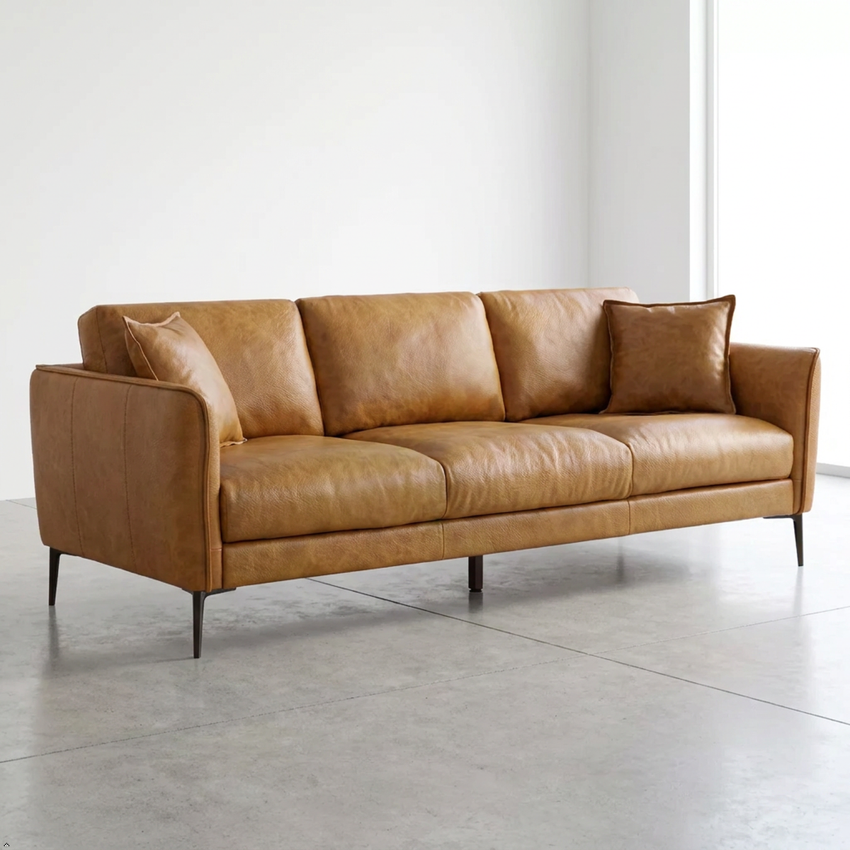 A mid-century modern three-seater sofa upholstered in warm tan leather, shown in a minimalist studio setting with a concrete floor and white walls.