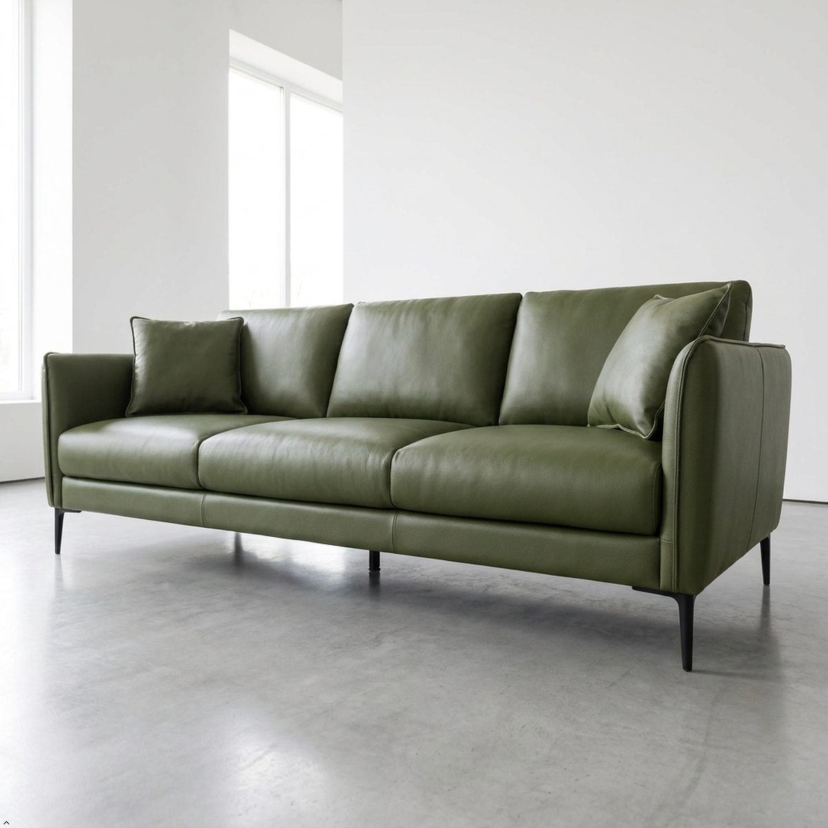 An olive green leather three-seater sofa with matching throw pillows, supported by slim black metal legs, set in a minimalist modern room with a concrete floor and white walls.