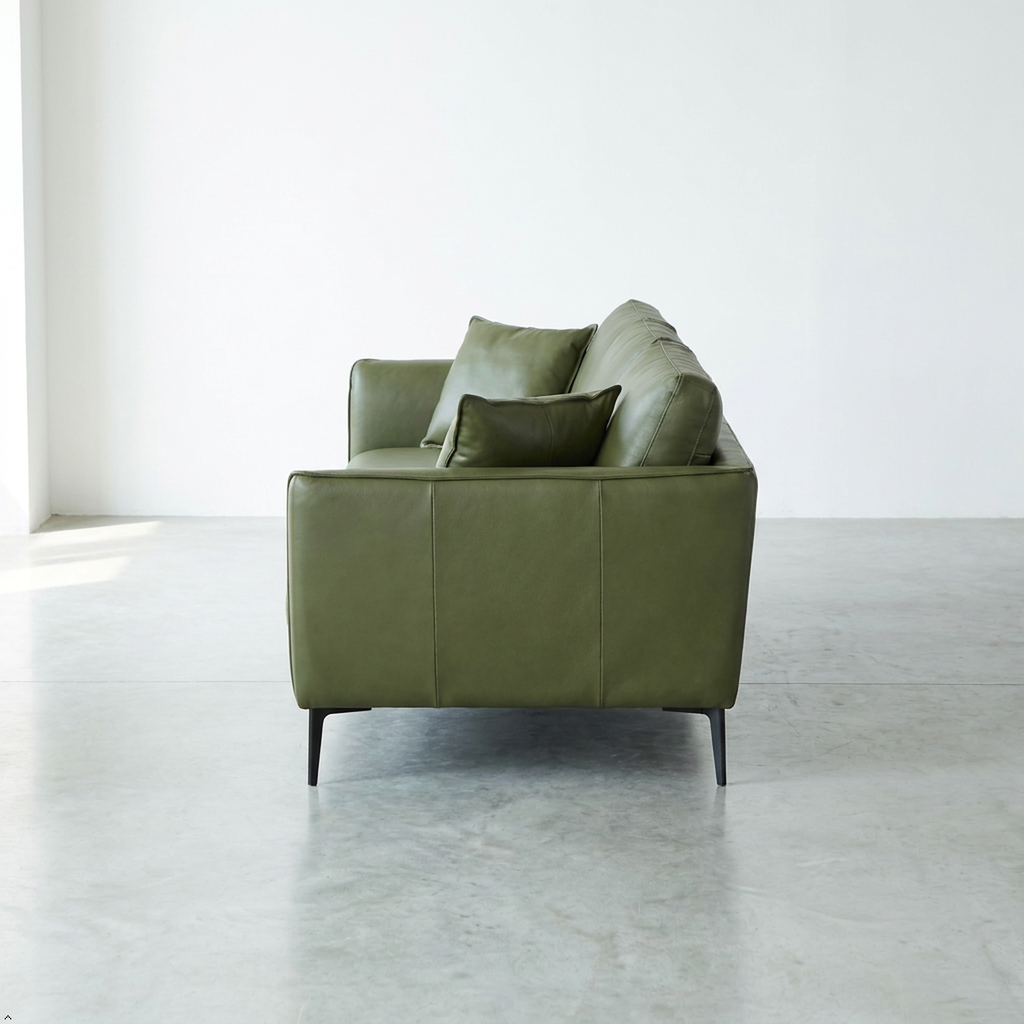 An olive green leather loveseat sofa with two matching throw pillows, positioned in a minimalist room with a concrete floor and white walls.