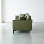 An olive green leather loveseat sofa with two matching throw pillows, positioned in a minimalist room with a concrete floor and white walls.