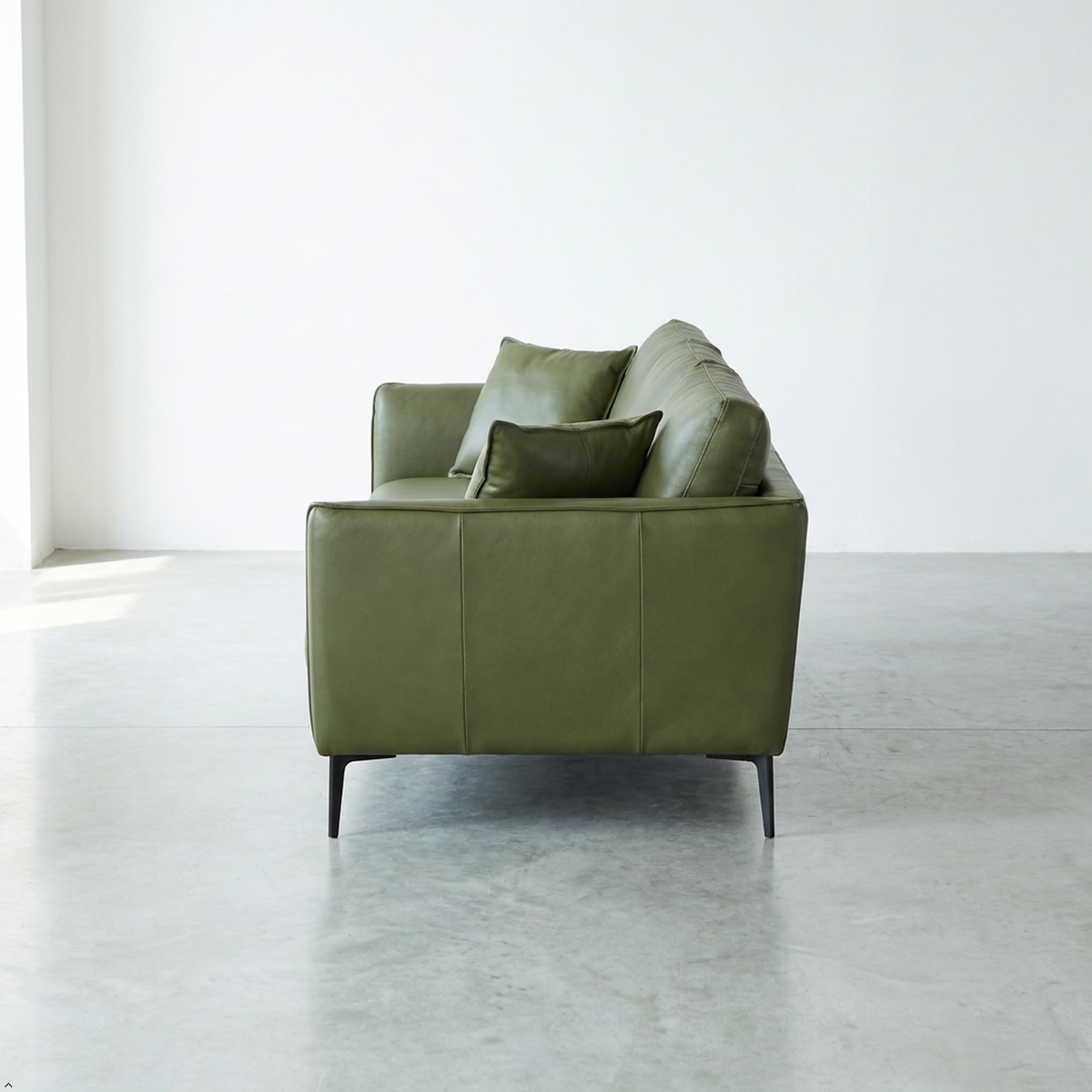 An olive green leather loveseat sofa with two matching throw pillows, positioned in a minimalist room with a concrete floor and white walls.