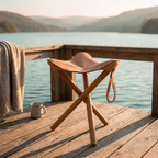 A tan leather and wood folding stool sits on a wooden deck overlooking a calm lake at sunset, with a knitted blanket draped over the railing and a mug nearby.