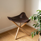 A dark brown leather folding stool with a natural wood X-frame base, set on a light wooden floor against a beige wall with a potted plant in the foreground.