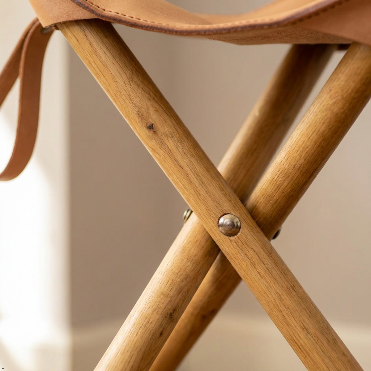 Close-up of a wooden folding chair with a tan leather seat and strap, showing the crossed legs and metal rivet detail.