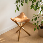 A modern folding stool with a tan leather seat and light wood legs, styled in a bright room next to a potted plant.