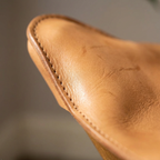 Close-up of a tan leather item, showing the texture and stitching along a curved edge against a blurred gray background.