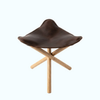 A modern stool with a dark brown leather seat and a light natural wood X-frame base, photographed on a seamless white background.