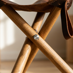 Close-up detail of a folding camp chair with a natural wood frame, brown leather seat and strap, and a silver metal rivet.