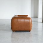 A modern low-profile leather armchair in a warm cognac brown, photographed from the side in a minimalist studio setting with a concrete floor and white walls.