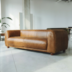 A brown leather sofa with rounded arms and wooden feet, positioned in a minimalist, modern room with concrete floors and light gray walls.