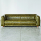A vintage-style three-seater sofa upholstered in distressed olive green leather, featuring rounded arms, channel tufting on the backrest, and small round wooden feet, set against a plain light gray wall and concrete floor.