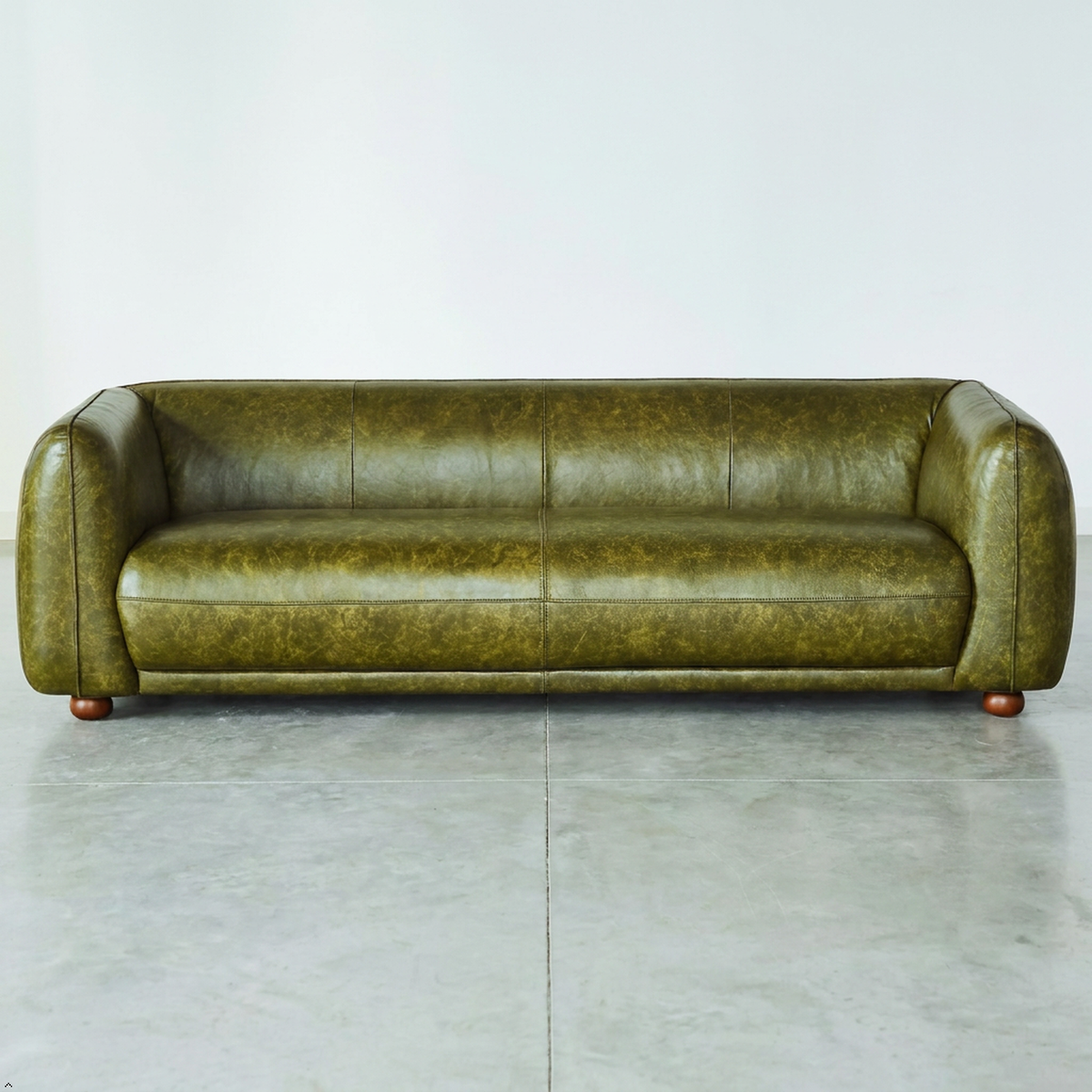 A vintage-style three-seater sofa upholstered in distressed olive green leather, featuring rounded arms, channel tufting on the backrest, and small round wooden feet, set against a plain light gray wall and concrete floor.