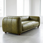 An olive green distressed leather loveseat sofa with a rounded, barrel-like silhouette and dark wooden feet, photographed in a bright, modern room with high windows and a concrete floor.