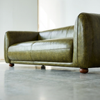 An olive green leather sofa with rounded arms and dark wooden ball feet, photographed in a minimalist room with a polished concrete floor.