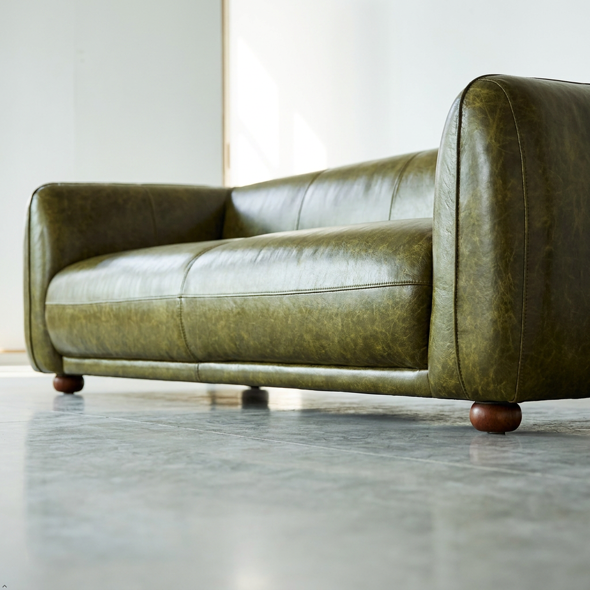 An olive green leather sofa with rounded arms and dark wooden ball feet, photographed in a minimalist room with a polished concrete floor.