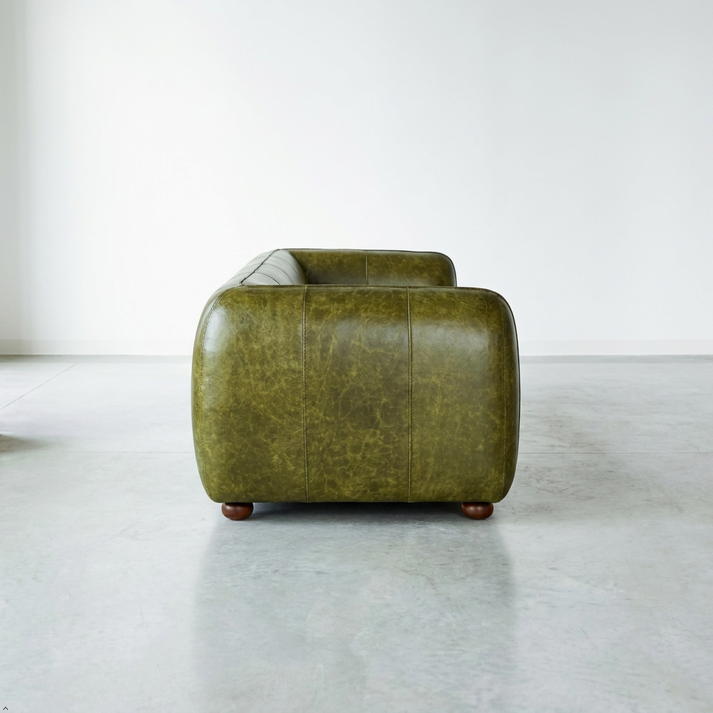 A low, wide armchair upholstered in distressed olive green leather with rounded blocky arms and small dark wooden feet, photographed in a minimalist studio setting with a light gray floor and white wall.