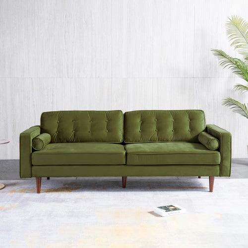 An olive green tufted velvet loveseat sofa with tapered wooden legs, positioned against a light gray textured wall with a potted palm in the background.