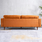 A mid-century modern two-seater sofa upholstered in a warm camel-brown velvet fabric, shown from the front against a light gray textured wall and concrete floor.