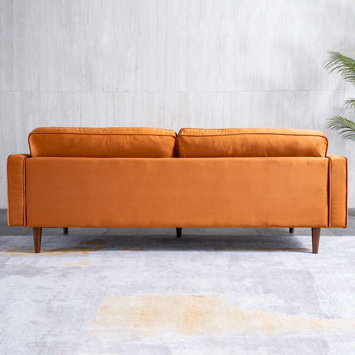 A mid-century modern two-seater sofa upholstered in a warm camel-brown velvet fabric, shown from the front against a light gray textured wall and concrete floor.