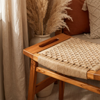 A wooden bench with a woven rope seat and a cutout handle on the side, styled in a warm, boho-lifestyle setting with pampas grass, curtains, and cushions.