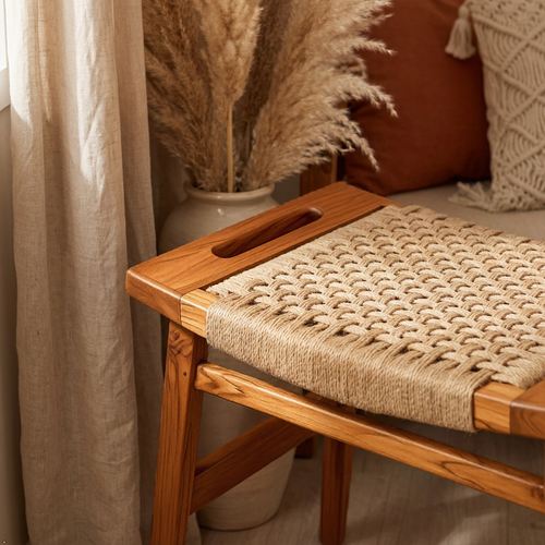 A wooden bench with a woven rope seat and a cutout handle on the side, styled in a warm, boho-lifestyle setting with pampas grass, curtains, and cushions.