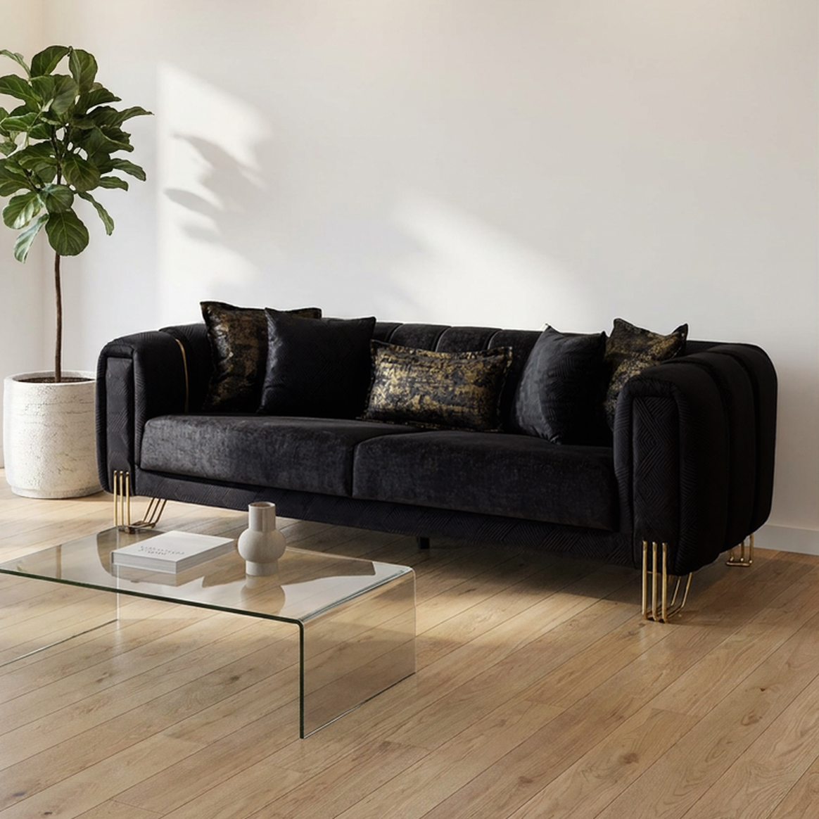 A modern black velvet sofa with gold metal legs and decorative throw pillows, set in a bright living room with light wood floors, a glass coffee table, and a potted plant.