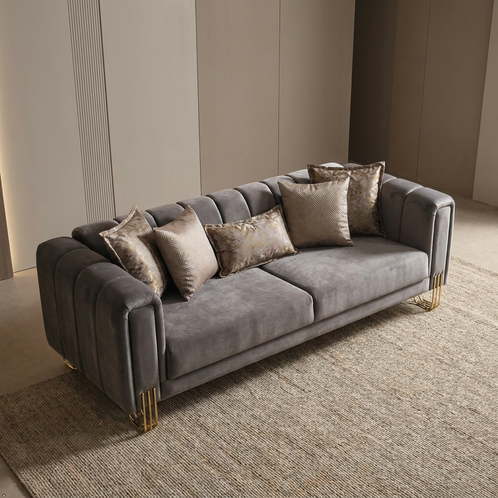A modern gray velvet sofa with channel tufting, styled with five textured beige throw pillows and gold metal legs, set on a woven rug in a minimalist beige room.