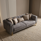 A modern gray velvet sofa with channel tufting, styled with five textured beige throw pillows and gold metal legs, set on a woven rug in a minimalist beige room.
