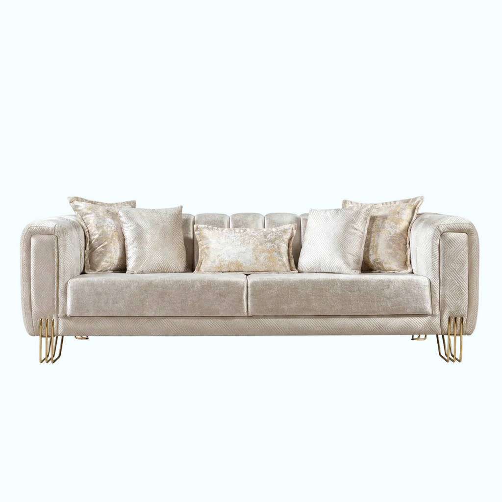 A modern three-seater sofa upholstered in a textured champagne beige fabric, styled with five matching throw pillows, including two with a shimmering gold pattern. The sofa features elegant curved arms and slim gold metal legs, set against a solid light blue background.