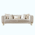 A modern three-seater sofa upholstered in a textured champagne beige fabric, styled with five matching throw pillows, including two with a shimmering gold pattern. The sofa features elegant curved arms and slim gold metal legs, set against a solid light blue background.