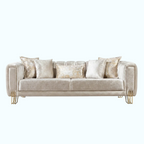 A modern three-seater sofa upholstered in a textured champagne beige fabric, styled with five matching throw pillows, including two with a shimmering gold pattern. The sofa features elegant curved arms and slim gold metal legs, set against a solid light blue background.