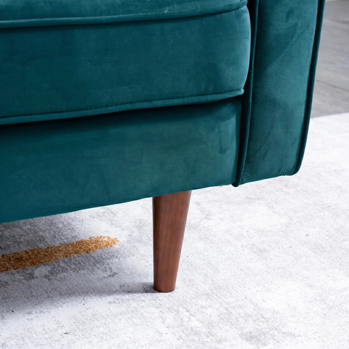 Close-up of a teal velvet upholstered chair or sofa corner with a tapered wooden leg, shown on a light gray rug.